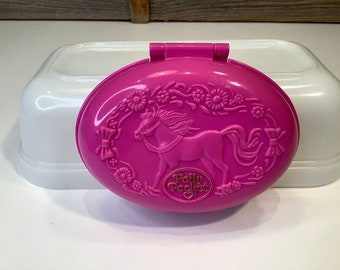 Op de afbeelding: Een roze Polly Pocket-compact met een witte basis. Het compact heeft een reliëfdesign van een paard met een bloemige rand en de tekst "Polly Pocket" in goud.