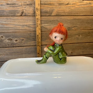 Peut inclure: Figurine vintage en céramique d'un elfe assis, avec des cheveux roux et une tenue verte. L'elfe est assis sur une surface blanche, avec une règle en bois en arrière-plan. La figurine a un design fantaisiste et caricatural.