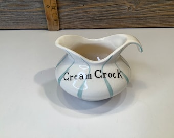 May include: A white ceramic creamer with a blue and white striped design. The creamer has a handle and says "Cream Crock" on the front.