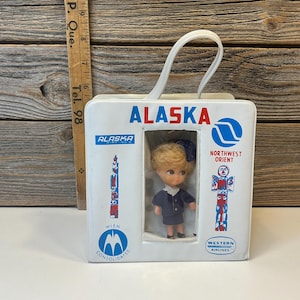 May include: Vintage Alaska Airlines doll in a white box with a handle. The box features the Alaska Airlines logo, the Northwest Orient logo, and the text "ALASKA". The doll is wearing a blue uniform.