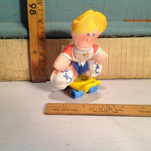 May include: A plush toy boy with blonde hair wearing a red, white and blue shirt and blue shorts is riding a yellow and blue toy skateboard. The toy is approximately 10 cm tall.
