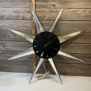 Puede incluir: Un reloj vintage con forma de estrella, con una esfera negra y rayos plateados. El reloj tiene un cable y está montado sobre una superficie de madera. El diseño del reloj recuerda al estilo moderno de mediados de siglo.
