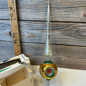 May include: A vintage glass Christmas tree topper featuring a tall, slender silver spire atop a gold spherical base with a green and red indented reflector centre. A wooden ruler shows the object stands approximately 29 cm in total height.