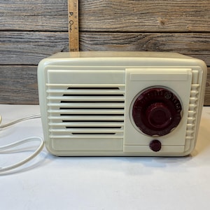 Puede incluir: Una radio rectangular de color crema con una rejilla de altavoz de listones horizontales a la izquierda y un dial de sintonización circular de color granate oscuro a la derecha. Un cable de alimentación blanco está unido al lado.
