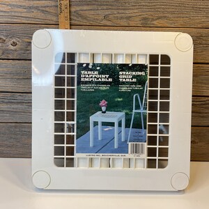 Peut inclure: Table en plastique blanc à grille empilable. La table est faite d'une grille en plastique blanc et est conçue pour s'empiler sur d'autres tables. La table est présentée sur une photo avec une petite table blanche et des chaises en arrière-plan. Le texte sur l'emballage indique "Table d'Appoint Empilable" et "Stacking Grid Table".