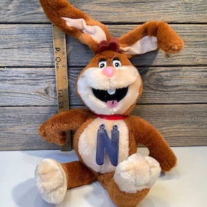 Puede incluir: Un conejito de peluche marrón con una gran sonrisa y una letra N azul en el pecho. El conejito está sentado sobre una superficie de madera.