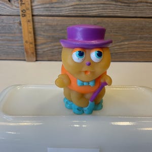 Puede incluir: Un juguete de goma amarillo con un sombrero morado, una camisa naranja y zapatos azules. El juguete sostiene un bast&oacute;n morado.