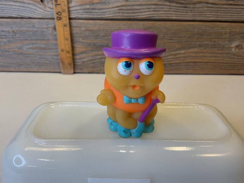 Puede incluir: Un juguete de goma amarillo con un sombrero morado, una camisa naranja y zapatos azules. El juguete sostiene un bast&oacute;n morado.