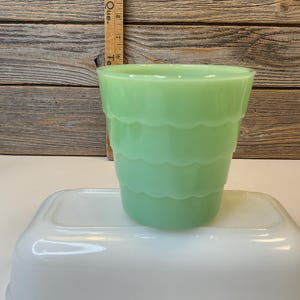 Peut inclure: Une tasse en verre jadeite vert clair présentant un design extérieur étagé et festonné. La tasse est placée devant une règle en bois affichant des mesures en centimètres et repose sur un plat rectangulaire en verre blanc.