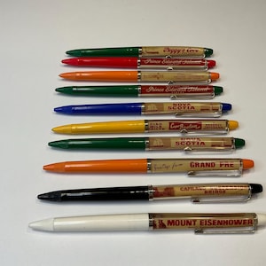 Op de afbeelding: Een verzameling kleurrijke pennen met verschillende bestemmingsnamen, waaronder "Peggy's Cove" en "Mount Eisenhower". De pennen zijn in groen, rood, oranje, blauw, geel, zwart en wit.