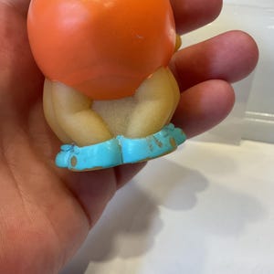 Puede incluir: Un peque&ntilde;o juguete de goma naranja con cabeza redonda y zapatos azules. El juguete est&aacute; sentado en una mano.