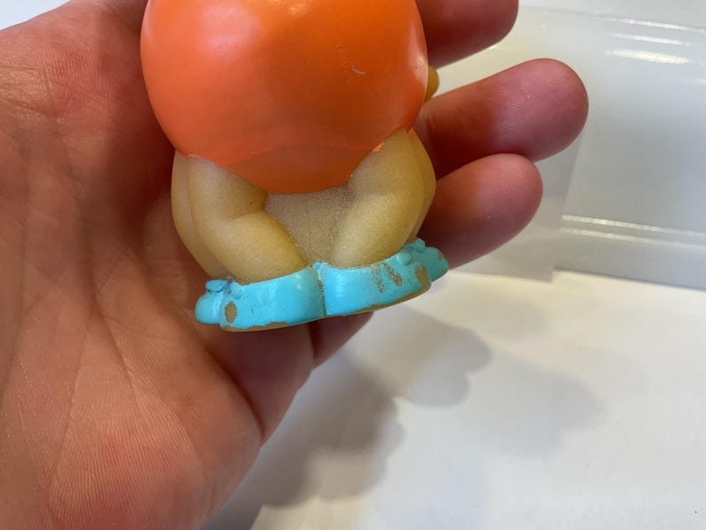 Puede incluir: Un peque&ntilde;o juguete de goma naranja con cabeza redonda y zapatos azules. El juguete est&aacute; sentado en una mano.