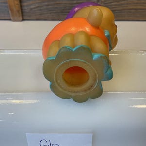 Puede incluir: Un juguete de goma colorido con un dise&ntilde;o morado, naranja y azul. El juguete tiene un agujero en la parte inferior y tiene forma de persona.