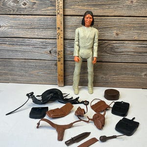 Peut inclure: Une figurine en plastique aux cheveux foncés portant une tenue à franges de couleur claire, placée à côté d'une règle en bois mesurant 40 cm. Divers accessoires en plastique marron et noir sont inclus, dont une selle, une tête de bison et des outils.