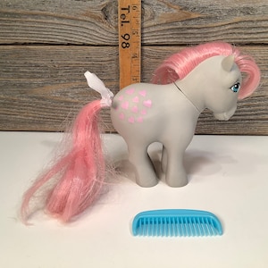 May include: A gray My Little Pony with pink hair and a white wing. The pony has pink hearts on its flank and is holding a blue comb.
