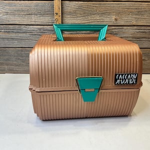 May include: A metallic rose gold plastic storage case with a ribbed texture, featuring a teal handle and a matching teal latch on the front. A black rectangular label with the white text SASSABY is attached to the right side of the case.