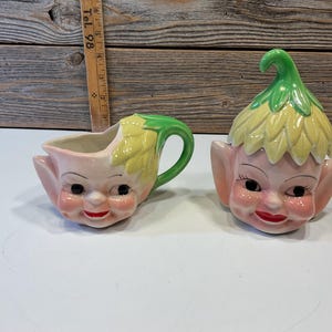 May include: A vintage ceramic creamer and sugar set featuring elf faces with green and yellow hats. The creamer has a handle and the sugar bowl has a lid.