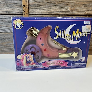 Peut inclure: Une boîte de jouet Sailor Moon Moon Cycle. La boîte est bleue avec le logo Sailor Moon et une image du jouet. Le jouet est rose, doré et violet, avec un design de croissant de lune et une étoile.