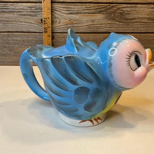 Può includere: Una teiera in ceramica a forma di uccellino blu con il viso rosa, grandi occhi con lunghe ciglia e un becco giallo. L'uccello presenta dettagli delle ali in blu scuro e un manico sul retro. Un righello in legno è visibile sullo sfondo.