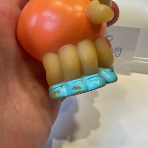 Puede incluir: Juguete de goma naranja con piernas amarillas y pies azules. El juguete tiene un cuerpo redondo y una cabeza peque&ntilde;a y puntiaguda.