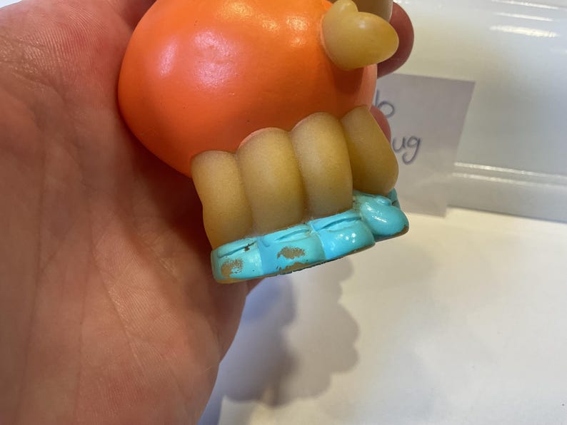 Puede incluir: Juguete de goma naranja con piernas amarillas y pies azules. El juguete tiene un cuerpo redondo y una cabeza peque&ntilde;a y puntiaguda.