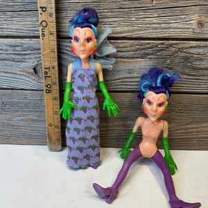 Peut inclure: Deux poupées aux cheveux violets et bleus, gants verts et boucles d'oreilles fraises. La plus grande porte une robe violette à motifs de raisins. La plus petite porte des leggings et bottes violets. Une règle en bois indique 28 cm de hauteur.