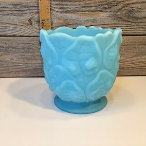 Puede incluir: Un jarrón de vidrio azul claro con un diseño floral texturizado. El jarrón tiene una boca ancha y una base redonda.