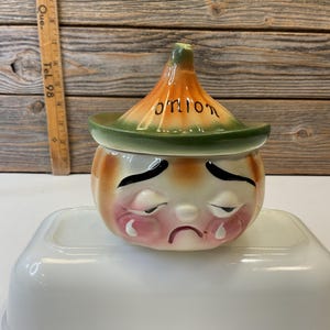 May include: A ceramic onion-shaped kitchen container with a green and orange lid. The lid has the word "ONION" written on it. The container has a sad face with tear-shaped drops of paint.
