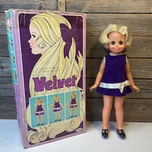 May include: A vintage doll with blonde hair and a purple velvet dress, next to its original box. The box features a side profile illustration of a woman with long blonde hair and the word "Velvet".