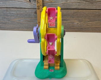 May include: A miniature Ferris wheel toy with a yellow wheel, pink seats, and a green base. The wheel has pink seats with star and heart details. The base is green with a purple handle. The toy is set on a white surface.