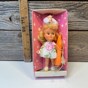 May include: A vintage doll with blonde hair and blue eyes, wearing a white dress with pink floral accents, is displayed in a pink box. The doll holds an orange accessory. The box has a white base and pink polka dots.