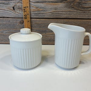 Peut inclure: Un sucrier en céramique blanc avec un couvercle et une crémière assortie. Les deux pièces présentent des détails verticaux côtelés. Le sucrier est rond et la crémière a une anse et un bec. Le fond est en bois.