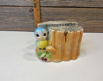May include: A ceramic planter shaped like a tree stump, featuring a bluebird perched on the side. The bird has a yellow belly and is surrounded by green leaves and red berries. The planter is painted in shades of brown, blue, and yellow.