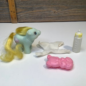 May include: A light blue pony with a yellow mane and tail, a white cloth nappy, a pink teddy bear, and a white bottle with a yellow lid.