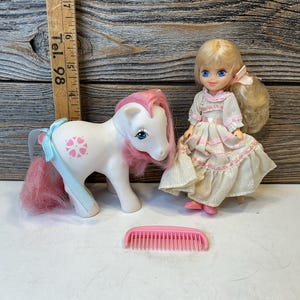 May include: A white toy pony with pink mane and tail, blue eyes, and a light blue ribbon. A doll with blonde hair and a pink dress stands next to the pony. A pink comb is in front of them.