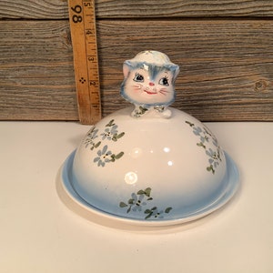 Puede incluir: Un mantequillero de cerámica blanca con un diseño floral azul y blanco. La tapa presenta una figurita de gato azul con cara blanca y ojos negros.