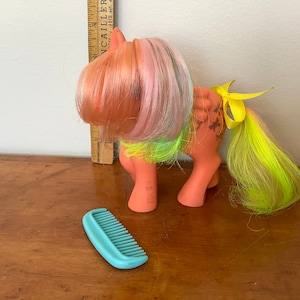 Puede incluir: Un juguete vintage de My Little Pony con pelo rosa y verde, una cinta amarilla y marcas marrones y negras. El pony está de pie sobre una superficie de madera con un peine azul.