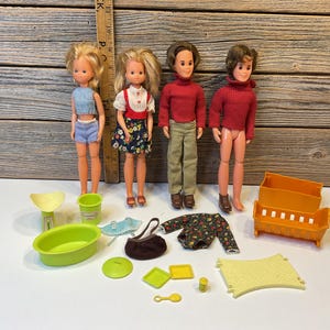 May include: A set of four vintage dolls, two girls and two boys, with accessories including a cot, a bath, a handbag, a blanket, and a plate. The dolls are dressed in colourful clothing, and the accessories are in various shades of green, yellow, and orange.