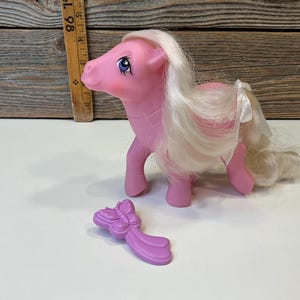 Peut inclure: Un poney rose avec des cheveux blancs et des yeux bleus. Un accessoire en forme de papillon violet est à côté du poney. Le poney a un ruban blanc noué autour de sa queue. Le fond est une surface en bois.