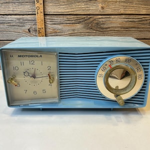 Könnte beinhalten: Ein hellblaues Motorola-Radiowecker im Vintage-Stil. Das Zifferblatt ist weiß mit schwarzen Ziffern und Zeigern. Auf der rechten Seite befindet sich ein Zifferblatt mit Temperaturangaben. Retro-Design.