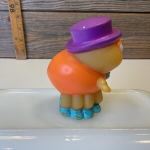 Puede incluir: Una peque&ntilde;a figura de juguete naranja y morada con zapatos azules. La figura lleva un sombrero morado y tiene una cara amigable.