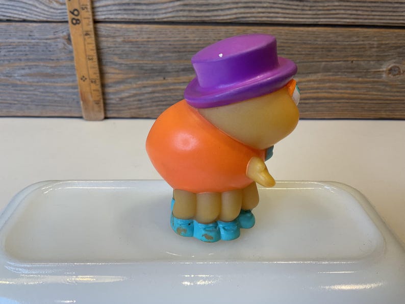 Puede incluir: Una peque&ntilde;a figura de juguete naranja y morada con zapatos azules. La figura lleva un sombrero morado y tiene una cara amigable.