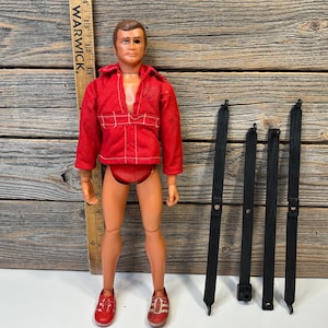 May include: A vintage action figure of a man in a red jacket, shorts, and trainers. The figure has brown hair and is next to black accessories and a wooden ruler. The ruler has the word "WARWICK" printed on it.