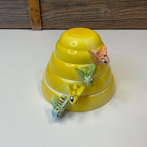 Op de afbeelding: Een gele keramische bijenkorfvormige container met vier decoratieve bijenbeeldjes. De container heeft een spiraalvormig ontwerp en is geschilderd in een heldere gele kleur. De bijen zijn in tinten roze, groen en geel.