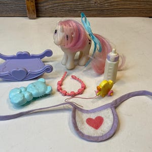 May include: A cream-coloured toy pony with pink hair and a blue ribbon, accompanied by a purple cradle, a light blue bear-shaped brush, a pink beaded necklace, a white baby bottle, a yellow duck toy, and a white bib with a red heart.