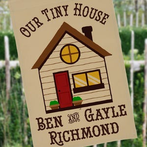 May include: A beige garden flag with a cartoon illustration of a brown house with a red door and yellow windows. The text "Our Tiny House" is at the top of the flag. The text "Ben and Gayle Richmond" is at the bottom of the flag.