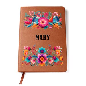 May include: A brown leather-bound journal with the name "MARY" in black block letters. The journal is decorated with colorful floral designs in pink, blue, orange, and purple. A matching ribbon bookmark is visible.