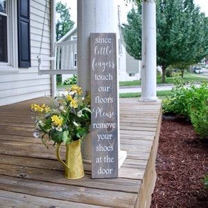 May include: A wooden sign with the text "Since little fingers touch our floors Please remove your shoes at the door" on a porch with a swing and a flower arrangement in a gold pitcher.