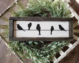 May include: A wooden sign with a black frame and a white background. Five black silhouettes of birds are perched on a wire. The sign is sitting on a white wicker basket filled with green foliage.