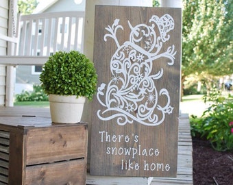 May include: A rectangular wooden sign with a dark brown stained finish featuring a white graphic of a snowman composed of intricate swirls and snowflakes. Below the snowman, the text reads, There's snowplace like home in white lettering.